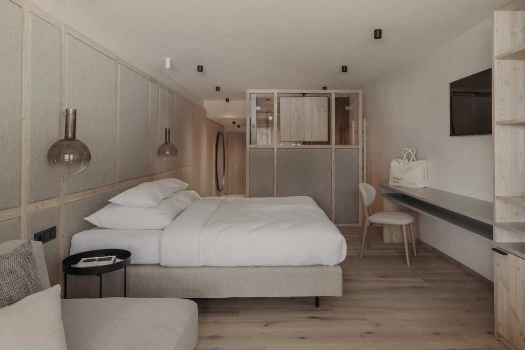 Hotel Marten Saalbach, Hotelzimmer, Boxspringbett vor tapezierter Rückwand, Scheibtisch mit Stuhl, Raumtrenner, Einrichtung in Lärche, Spiegel rund